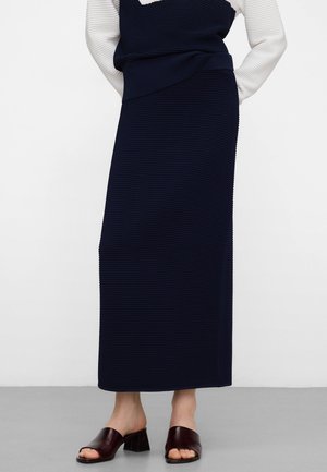 Marineblauer Maxirock mit Rippstruktur und einem strukturierten Bund, kombiniert mit braunen offenen Schuhen mit Blockabsatz. Glatte Texturen sichtbar.