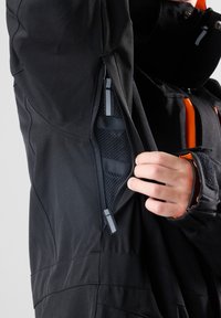 Schwarze Jacke mit einem Reißverschlusstaschen-Detail und einem Mesh-Futter. Der Stoff hat eine glatte Textur und ist funktional gestaltet.