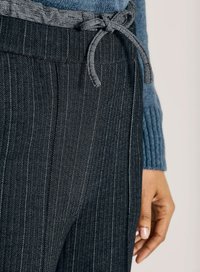 Close-up van donkere streeppantalon met een gestrikte stoffen riem, gedragen met een blauwe gebreide trui, waarbij een ontspannen hand aan de zijkant zichtbaar is.