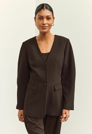 Frau mit zurückgebundenem Haar, die goldene Creolen trägt und einen dunkelbraunen, geknöpften Blazer über einer passenden Hose trägt, steht vor einem einfarbigen Hintergrund.