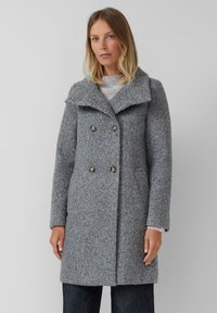 Vrouw in een grijze wollen dubbelbreasted jas met brede kraag en knoopdetails, gecombineerd met donkere broek tegen een effen achtergrond.