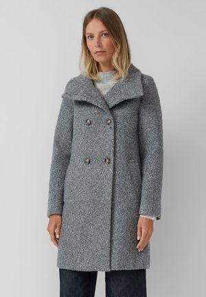 Femme portant un manteau gris en laine à double boutonnage avec un large col et des détails de boutons, associé à un pantalon sombre sur un fond uni.
