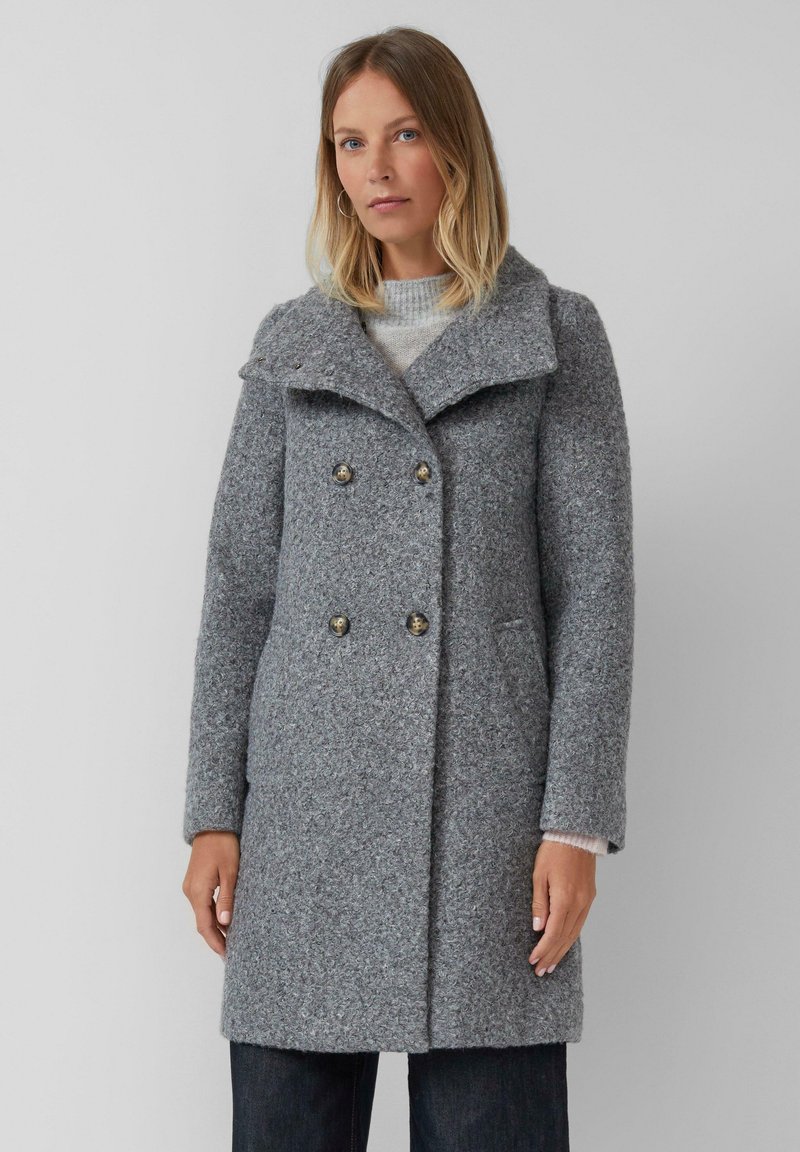 Vrouw in een grijze wollen dubbelbreasted jas met brede kraag en knoopdetails, gecombineerd met donkere broek tegen een effen achtergrond.
