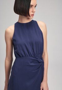 Sleeveless, navy blue dress with a relaxed fit, gathered waist, and a decorative knot detail at the side. Smooth, matte fabric texture.