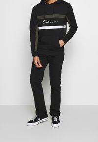 Sudadera con capucha negra con rayas verdes y blancas, que cuenta con un bolsillo frontal y un logo en escritura. Combinada con pantalones negros y zapatos negros con detalles en blanco.