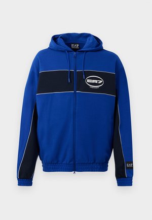 Plava zip-up dukserica s crnom horizontalnom prugom i bijelim logotipom, elastičnim manžetama i kapuljačom s pertlom. Mekani materijal s ležernim dizajnom.