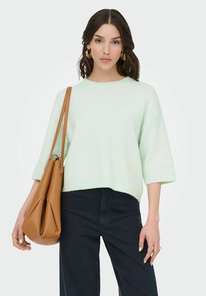 Femme aux cheveux foncés portant un pull vert clair, un pantalon sombre, de grandes boucles d'oreilles en or et un sac cabas en cuir souple marron sur l'épaule.