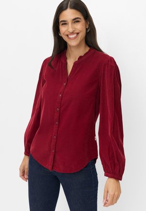 Kvinde med langt mørkt hår iført en rød knap-op bluse med puffede lange ærmer og mørkeblå jeans, smilende mod en hvid baggrund.
