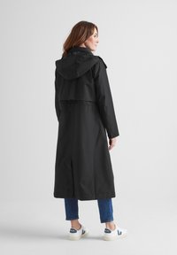 Black waterproof trench coat with a hood, long design, and adjustable waist. Worn over blue jeans and white sneakers with navy accents.