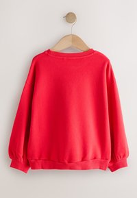 Felpa rossa con maniche lunghe e voluminose, collo e orlo a coste. Tessuto liscio e morbido, appesa a una gruccia di legno su uno sfondo chiaro.