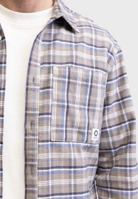 Kostkovaná flanelová košile v odstínech hnědé, modré a bílé. Obsahuje náprsní kapsu, zapínání na knoflíky a měkkou texturu. Nositá přes bílou košili.