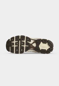 Semelle d'une basket montrant une bande de roulement noire et beige avec des motifs rectangulaires et courbes sur fond blanc.