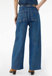 Laineet modaat siniset farkut leveillä lahkeilla ja korkealla vyötäröllä, joissa on kaksi takataskua ja raaka helmitys. Kangas on sileä tekstuuri.