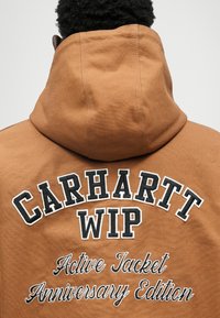 Καφέ φούτερ με μεγάλο παρα embroidered λογότυπο "CARHARTT WIP" και κείμενο "Active Jacket Anniversary Edition" σε λευκό και μαύρο.