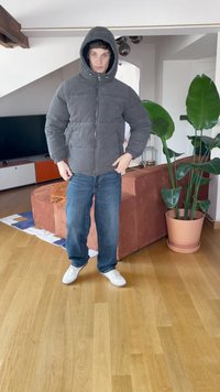 Casaco puffer cinza com capuz, fecho em zip, e punhos elásticos, combinado com jeans azuis e ténis brancos, em um ambiente interior iluminado.