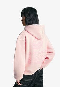 Sweatshirt roz cu glugă, cu o croială relaxată, având un print cu buze și textul 'expoziție în aer liber' pe spate. Fabricat dintr-un material moale și texturat.