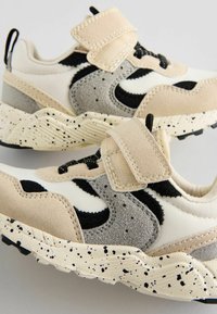 Baskets de sport beiges et grises avec strap en velcro, accents en daim texturé, détails noirs et semelle crémeuse mouchetée. Caractéristiques de design moderne.