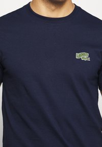 Marinblå bomullströja med rund krage. Har en liten grön och vit broderad Lacoste-logga på vänster bröst.