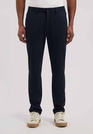 Uomo che indossa pantaloni slim fit blu navy e sneakers bianche, in piedi davanti a uno sfondo chiaro e uniforme.