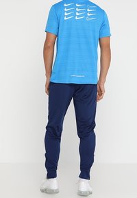 Blauw sportshirt met korte mouwen en witte Nike-logo's op de achterkant, gecombineerd met navy fitted broek en lichtgrijze sportschoenen.