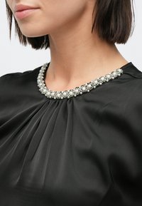 Μαύρο τοπ με πιέτες και στρογγυλή λαιμόκοψη διακοσμημένη με ασημένια και μαργαριταρένια διακόσμηση. Λεία υφή.