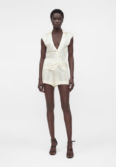 Γυναίκα που φοράει ένα κρεμ αμάνικο τοπ με V-λαιμόκοψη με κουμπιά και αντίστοιχα κοντά σορτς με δαντελένιες λεπτομέρειες, συνδυασμένο με μαύρα πέδιλα με λουράκια, όρθια μπροστά σε λευκό φόντο.