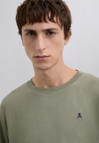 Camiseta de algodón color verde oliva con cuello redondo y mangas cortas. Presenta una pequeña calavera y huesos cruzados bordados en el pecho.