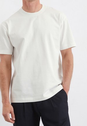 Man draagt een eenvoudige witte korte mouw T-shirt met ronde hals en zwarte broek, met één hand in de zak, staand tegen een witte achtergrond.