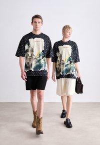 Sorte oversized t-shirts med et farverigt landskabsprint, parret med sorte og beige shorts. Tilbehøret inkluderer en brun taske og støvler.