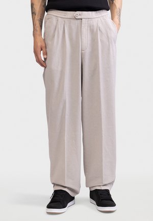 Άντρας που φοράει φαρδιά ανοιχτό μπεζ παντελόνια, μαύρα αθλητικά παπούτσια με λευκές σόλες, τα χέρια στις τσέπες, με ορατά τατουάζ στα μπράτσα, στέκεται σε λευκό φόντο.