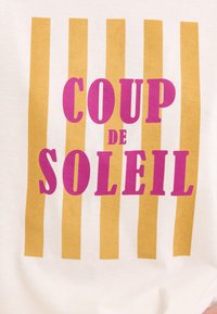 Tela blanca con rayas verticales amarillas. El texto central dice "COUP de SOLEIL" en letras gruesas de color rosa. Diseño simple con un aire juguetón.