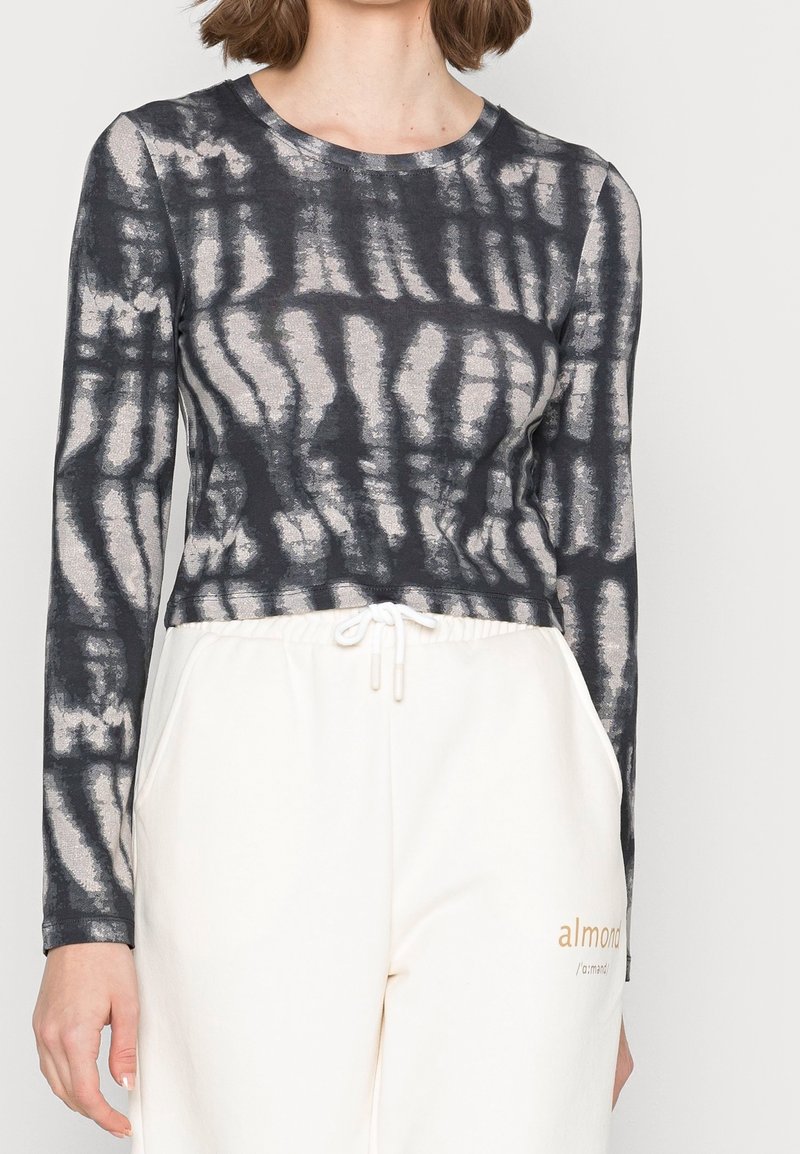 Maglia aderente a maniche lunghe di colore grigio scuro con motivo in tie-dye chiaro; tessuto morbido; scollatura rotonda; indossata con pantaloni della tuta chiari.