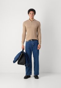 Maglione aderente color beige con zip e texture a coste; jeans a gamba larga blu; tiene una borsa nera e un giubbotto di jeans; scarpe nere robuste.