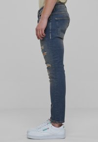 Peened blue denim püksid, millel on kulumisjäljed, madal talje, lõhikud ja nähtavad õmblused, koos valgete tossudega.