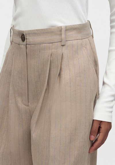 Pantalon beige à fines rayures taille haute avec plis, fermeture par boutons, passants pour ceinture et poches latérales, porté avec un haut blanc à manches longues.