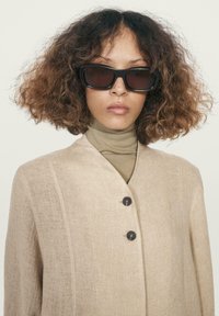 Kvinde med krøllet hår, iført store sorte solbriller, en beige knapsyet frakke og en lysebrun turtleneck mod en ensfarvet baggrund.