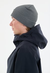 Grijze gebreide beanie gedragen met een marineblauwe hoodiejack. De beanie is aansluitend, met een gebreid patroon. De jas heeft een trekkoord bij de nek.