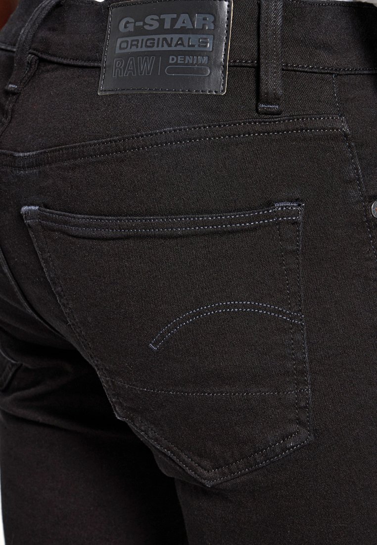 Des jeans en denim noir présentent une texture lisse, une étiquette en cuir à la taille et des coutures discrètes sur les poches arrière.