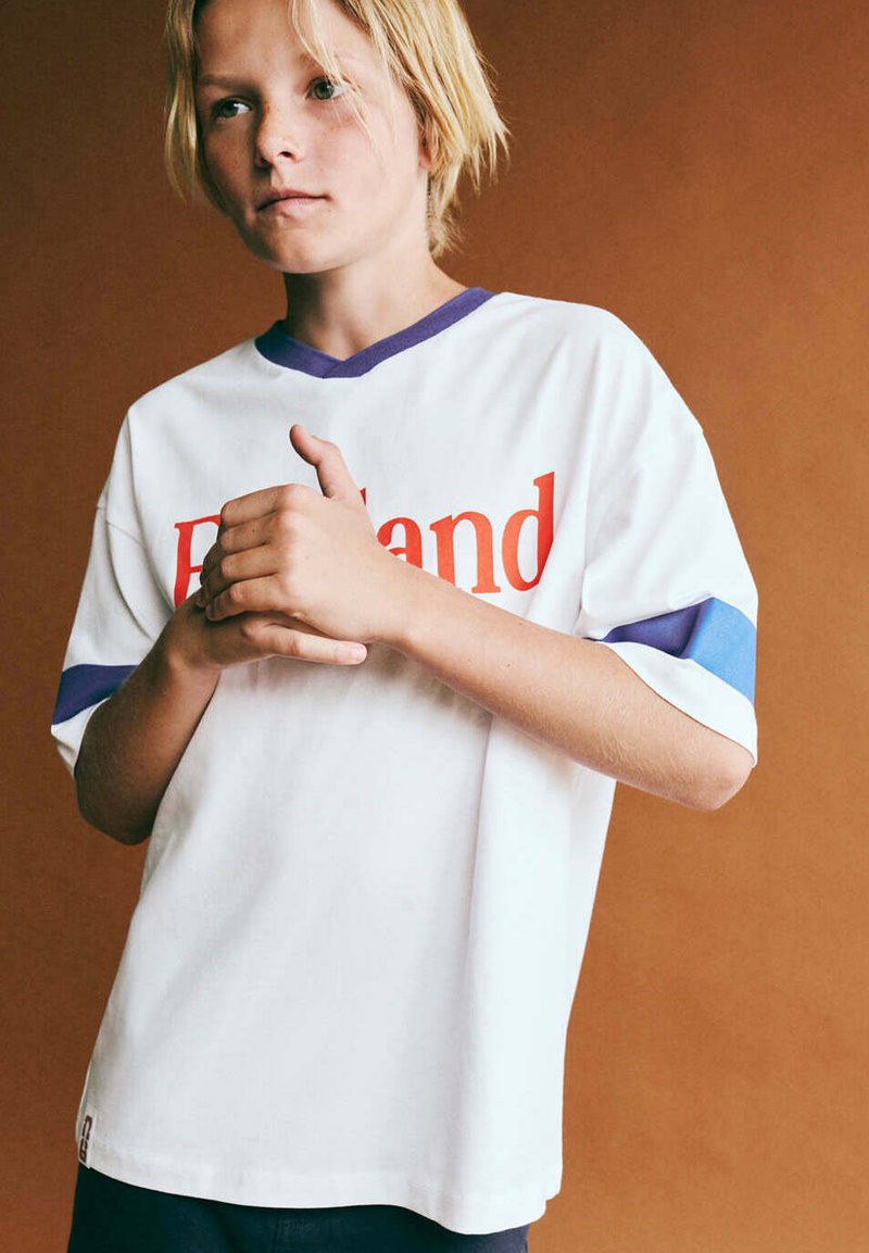 Enfant blond portant une chemise blanche avec un liseré bleu et un texte rouge, debout devant un fond marron uni, les mains jointes.