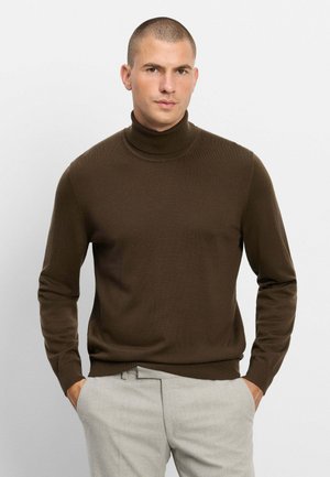 Uomo con capelli corti che indossa un maglione marrone a collo alto e pantaloni grigio chiaro con le mani nelle tasche, rivolto in avanti.