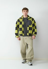 Vezelige geruite jas in limoen groen en zwart, met een ritszak in het zwart. Gecombineerd met losse beige broek en zilveren sneakers.