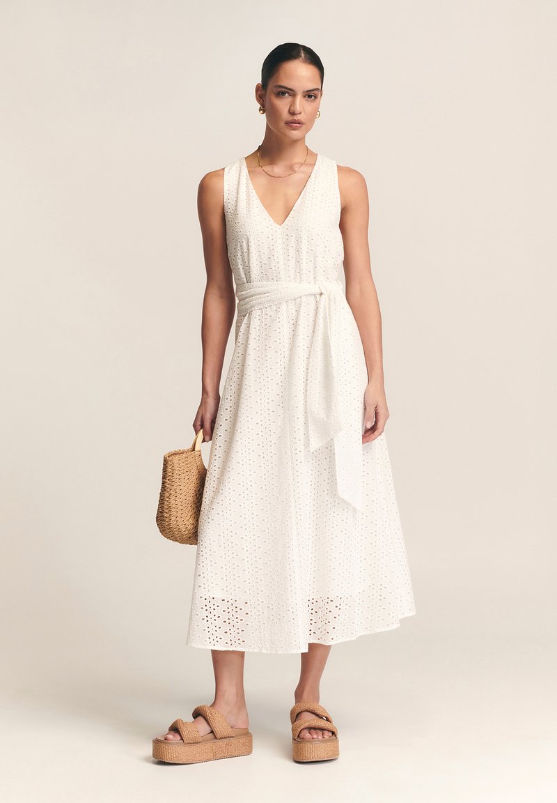 Balta akių maksi suknelė su V formos iškirpte, juosmens diržu ir laisva siluete. Derinama su rudos spalvos platformos sandaletėmis ir austine rankine.