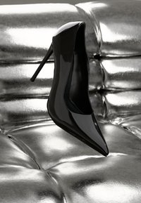 Black patent leather high heel with a pointed toe and a sleek design, resting on reflective silver upholstery.