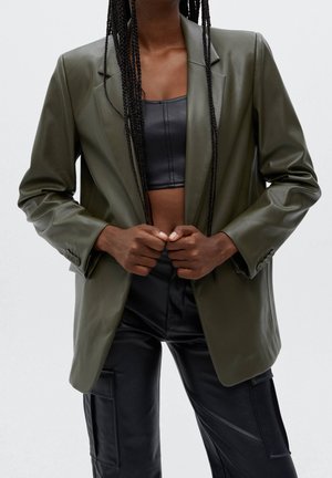 Personne avec de longues tresses portant un blazer en cuir vert olive sur un crop top noir et un pantalon cargo en cuir noir, ajustant l'avant du blazer.