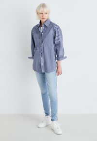 Chemise à rayures bleues et blanches avec les manches retroussées, associée à un jean skinny bleu clair et des baskets blanches sur un fond neutre.