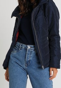Giacca imbottita navy con texture trapuntata, chiusura con zip e cappuccio. Indossata sopra una maglietta scura e abbinata a jeans blu chiaro a vita alta.