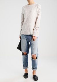 Lichtgrijze trui met een textuurafwerking, losse pasvorm en gestrikte mouwdetails; gecombineerd met versleten blauwe jeans en zwarte instap schoenen.