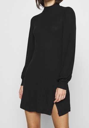 Kobieta ubrana w czarną dzianinową sukienkę z długim rękawem, stójką i bocznym rozporkiem, widoczna od szyi do połowy uda na jednolitym tle.