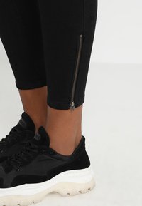 Jeans negros ajustados con cremalleras en los tobillos, combinados con zapatillas negras con parte superior texturizada y suela gruesa blanca.