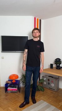 Μαύρη κοντομάνικη μπλούζα με κόκκινο κείμενο, μπλε τζιν, και σκούρες loafers σε δωμάτιο με μοντέρνα έπιπλα και εσωτερικά φυτά.